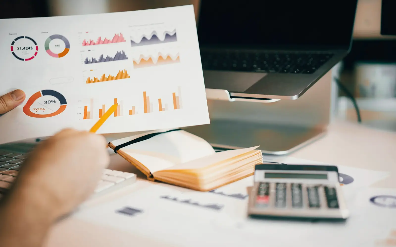 A hand holding a page of sales charts over a desk with a laptop and calculator