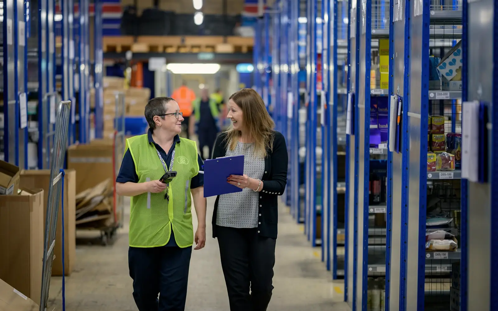 Two colleagues with a clipboard walking down a warehouse aisle of blue shelving