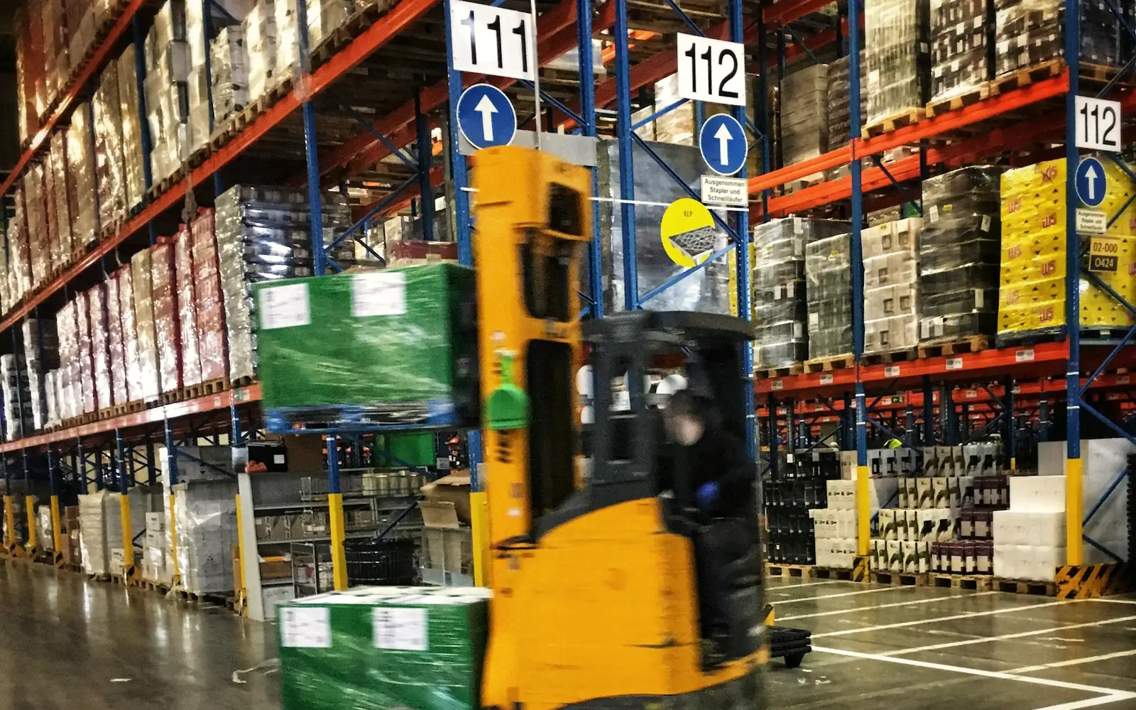 A forklift moving pallets of shrink-wrapped goods past tall warehouse racking