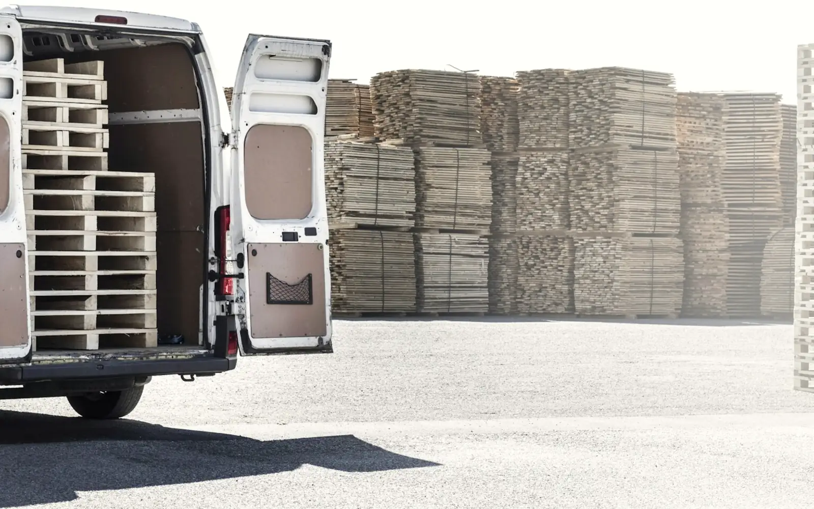 An open delivery van loaded with wooden pallets, parked beside stacks of pallets in a yard