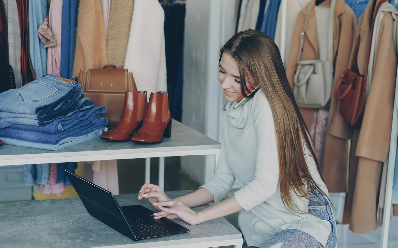A shop owner working on a laptop among racks of clothing